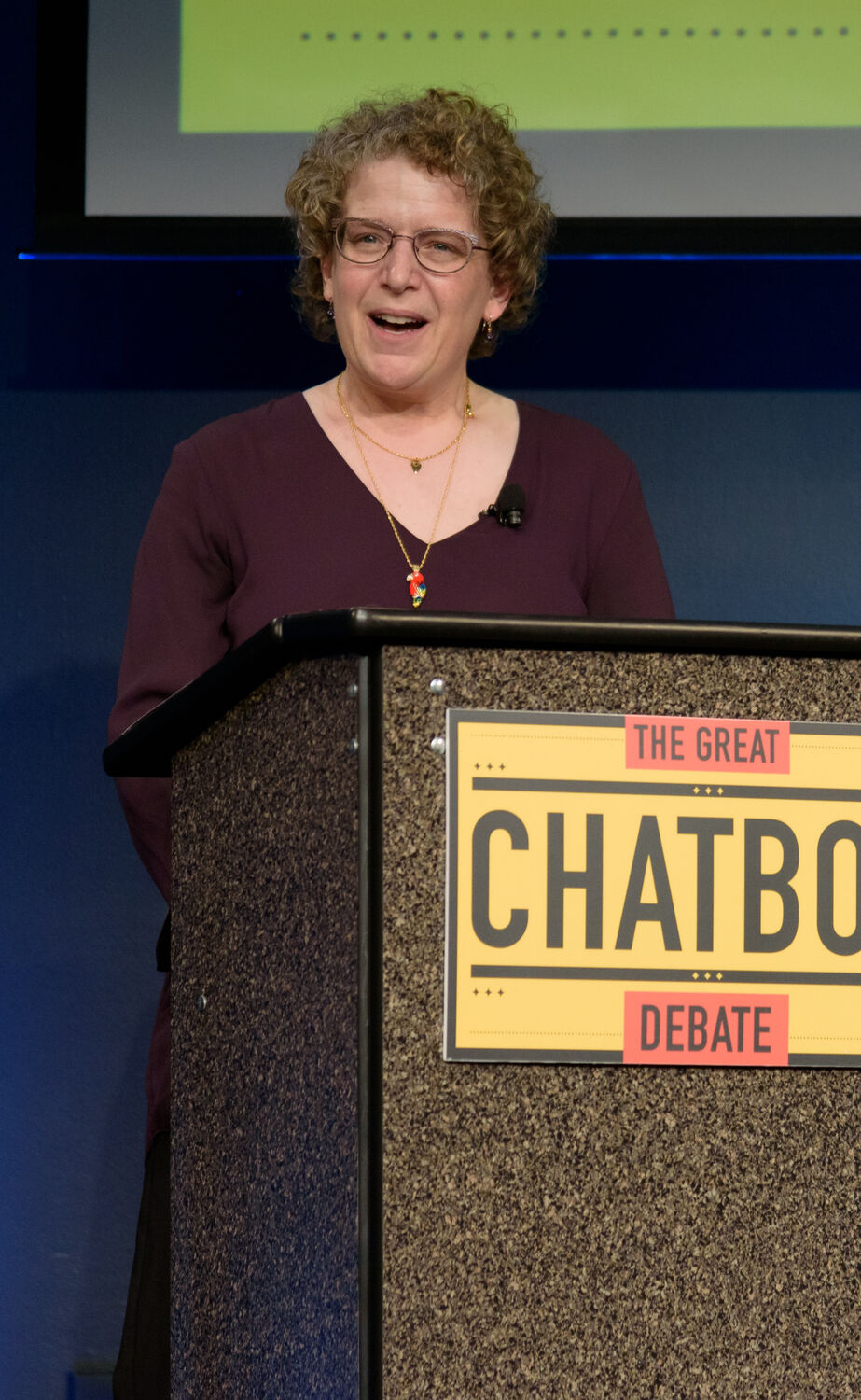 A woman with short curly hair and glasses, in a plum top, smiling as she speaks at a lectern whose sign reads 'The Great Chatbot Debate'