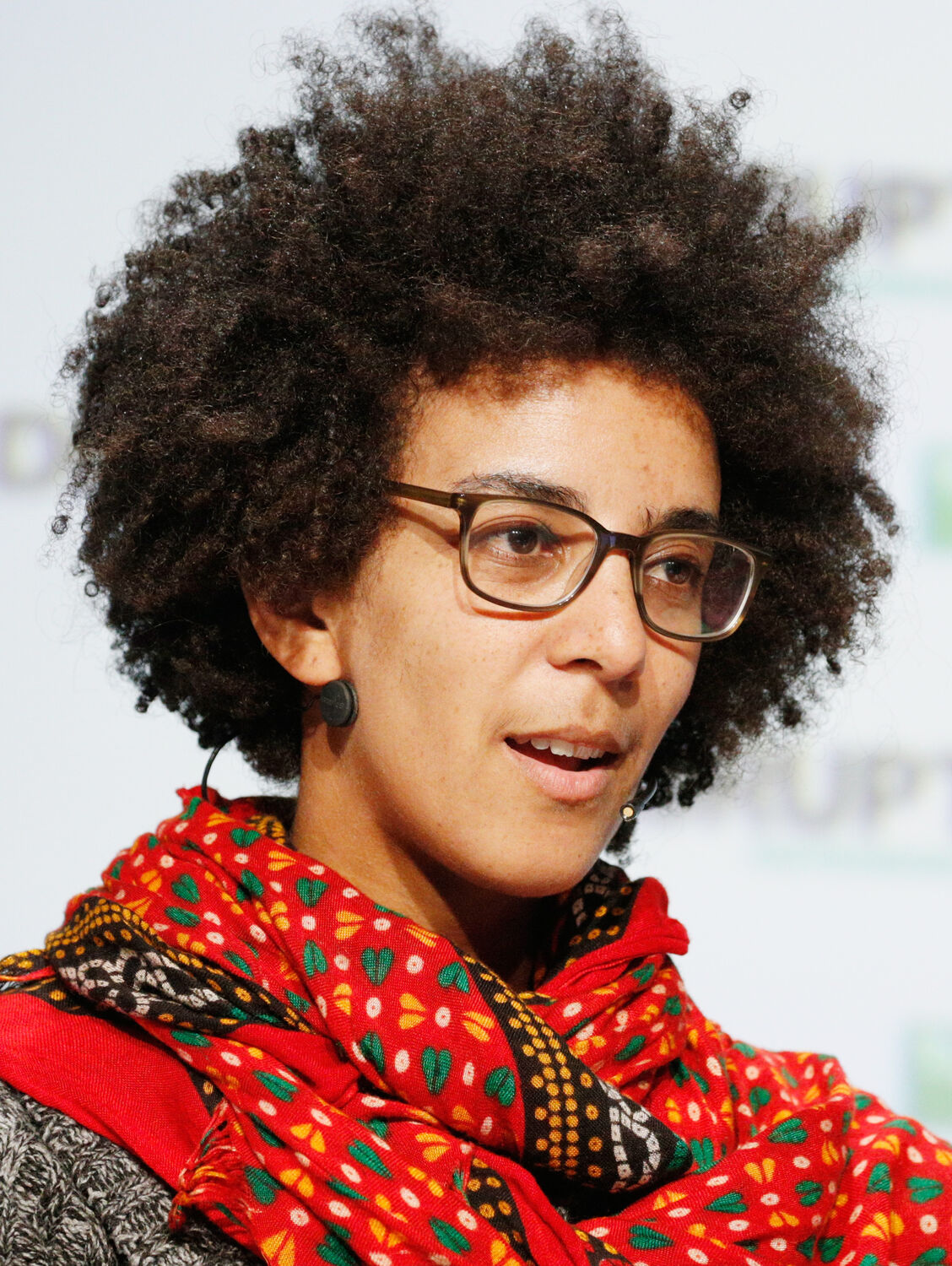 A woman with a large natural afro and dark-rimmed glasses, wearing a red patterned scarf, speaking on a stage