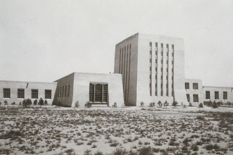 A tall stack tower with narrow vertical windows and low flat-roofed wings in Pueblo Revival style, standing alone behind open sand dotted with young shrubs and tumbleweeds