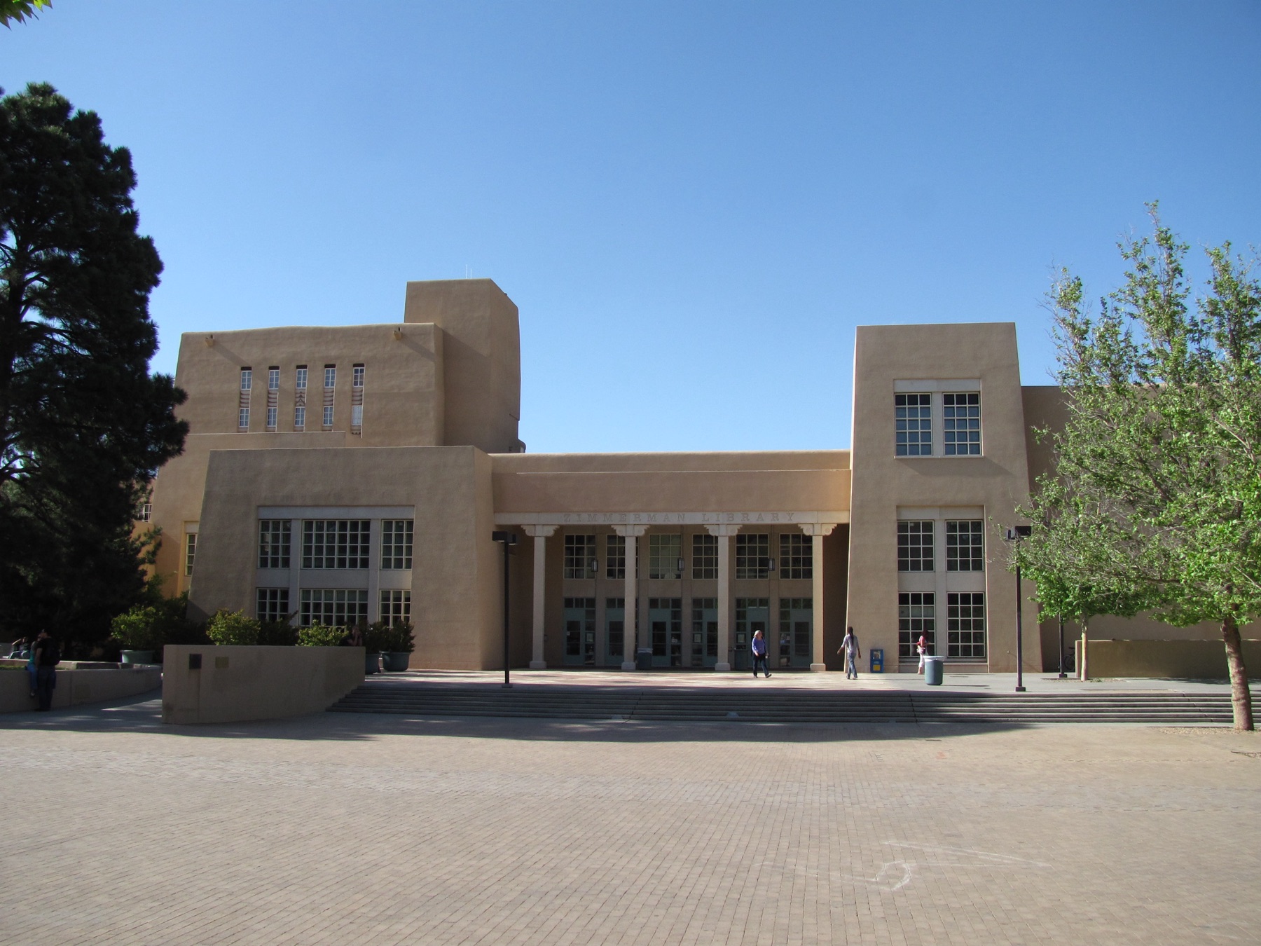 A large tan Pueblo Revival building with stepped, flat-roofed masses and a central tower, behind a lawn and trees under a blue sky