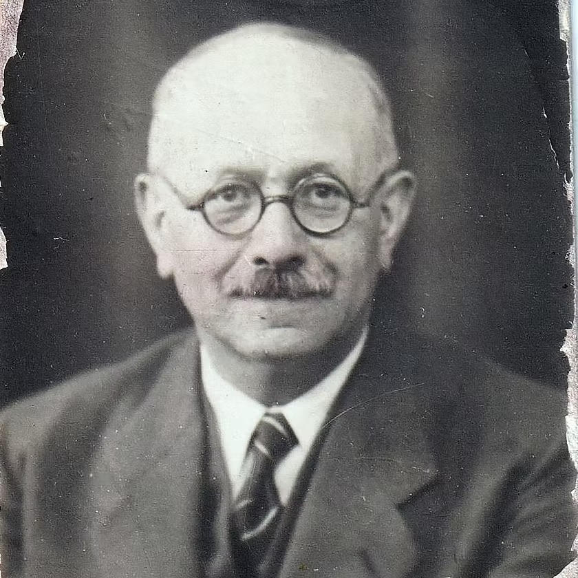 A worn identity photograph of a balding man in his forties with round wire spectacles, a clipped moustache, a dark suit and a striped tie, looking straight at the camera