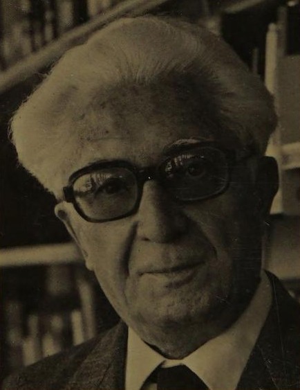 A sepia photograph of an elderly man with swept-back white hair and heavy dark-framed glasses, in a tweed jacket and tie, shelves of books behind him
