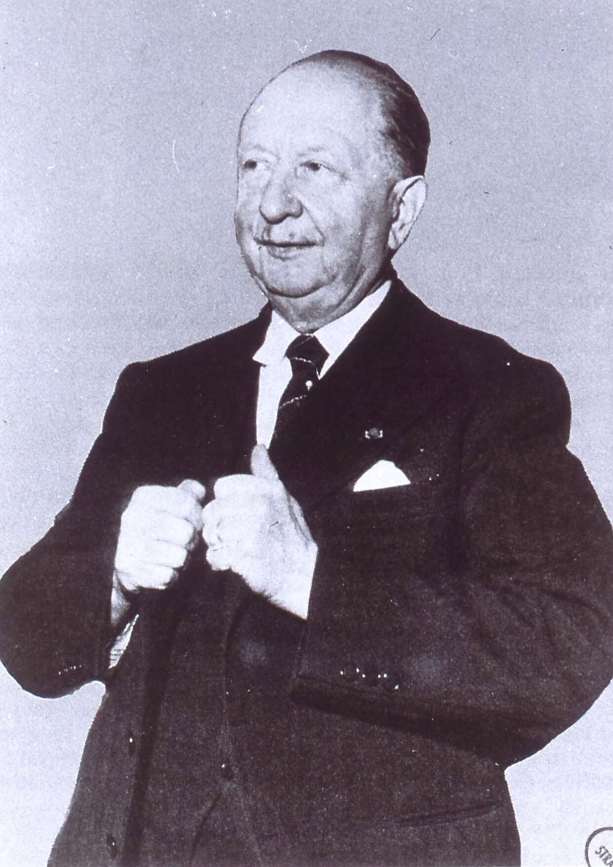 A press photograph of an elderly bald man in a dark three-piece suit, half-smiling, both gloved hands resting on the handle of a walking stick, a rosette in his lapel