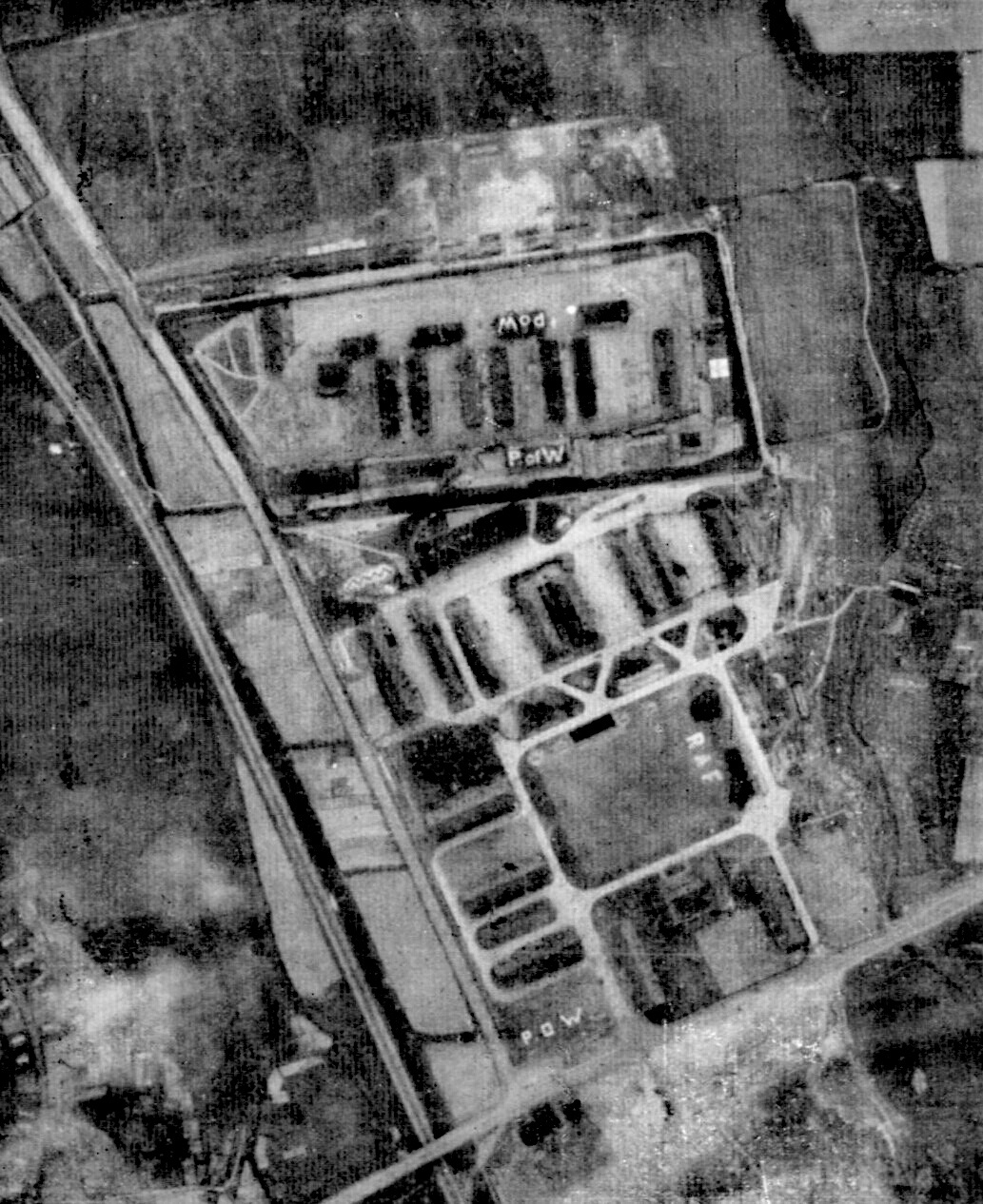 A grainy black-and-white aerial photograph of a prison camp: rows of long huts inside wire-fenced compounds beside a road and a rail line, with the letters P.o.W and R.A.F laid out in white on the open ground