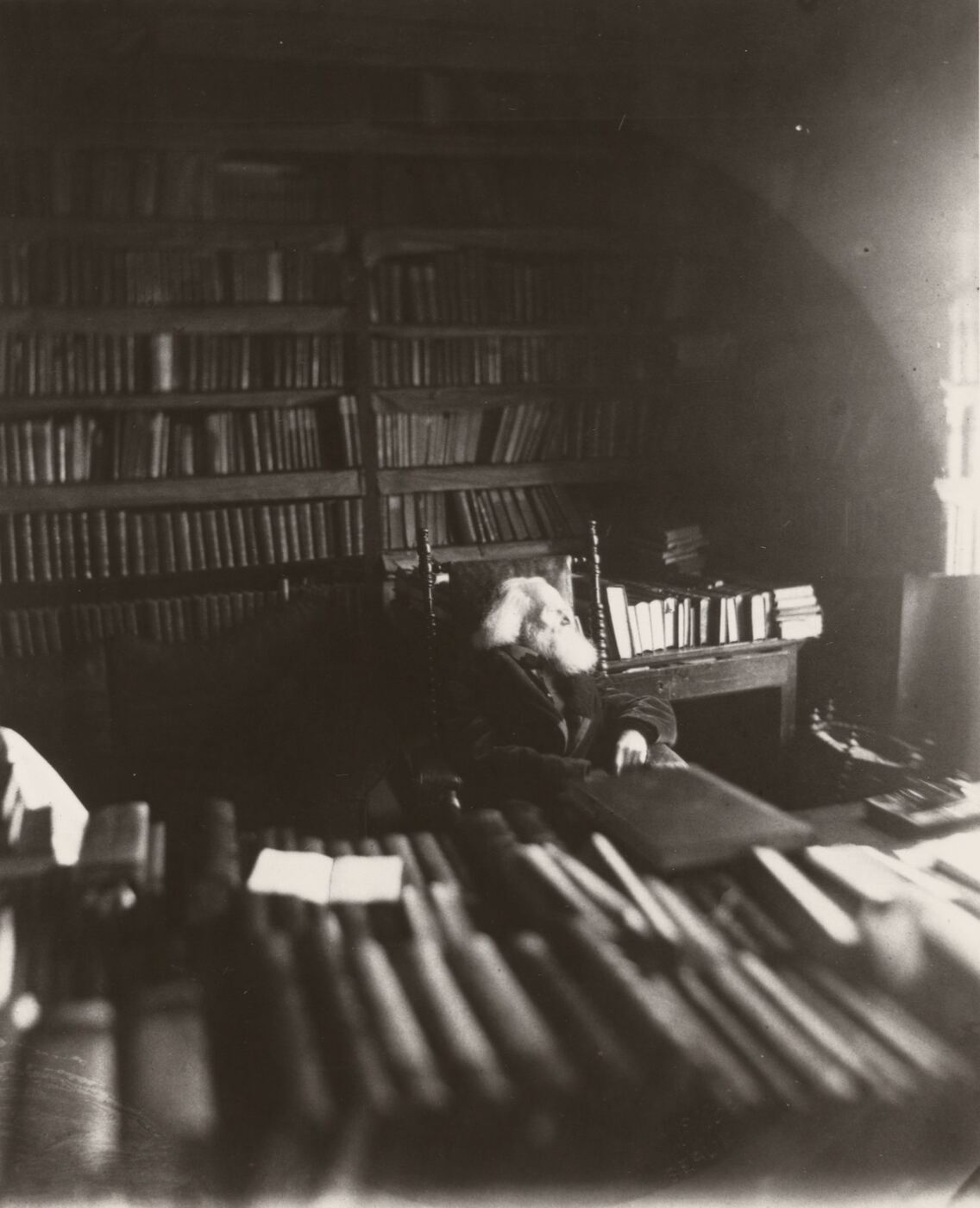 A dim photograph of a room walled with bookshelves, papers stacked in front, an old man barely visible at a desk