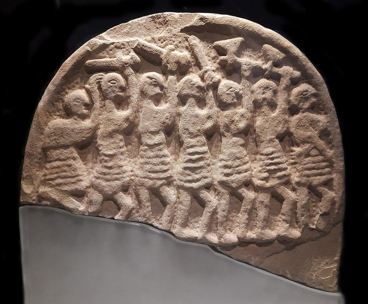 One face of a carved sandstone grave marker: seven armed figures in a row raising swords and axes