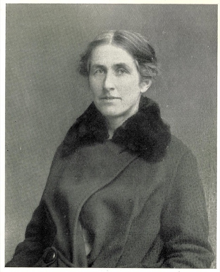 A black and white studio photograph of a woman in late middle age in a dark fur-collared coat, hair drawn back, looking past the camera