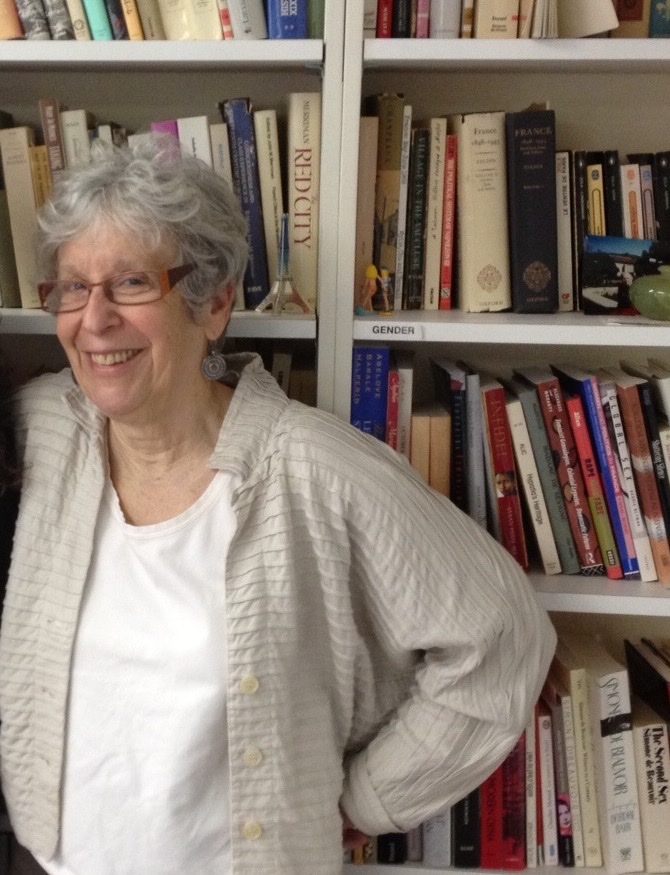 A colour photograph of an older woman with short white hair and red-framed glasses, smiling, standing in front of crowded white bookshelves; one shelf carries a label reading GENDER
