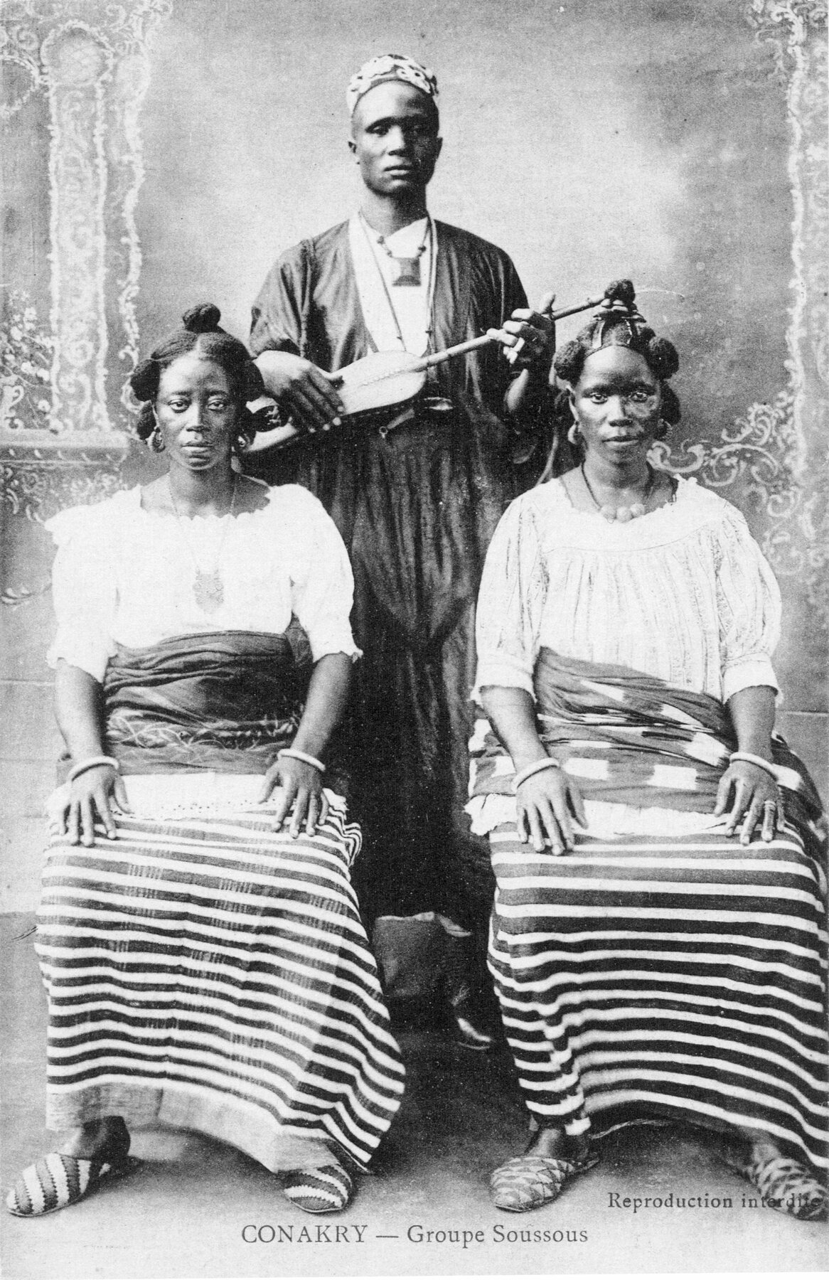 A studio photograph printed as a postcard: a standing man in a robe and cap holding a small lute, two seated women in white blouses and striped wrappers in front of him, a painted backdrop behind