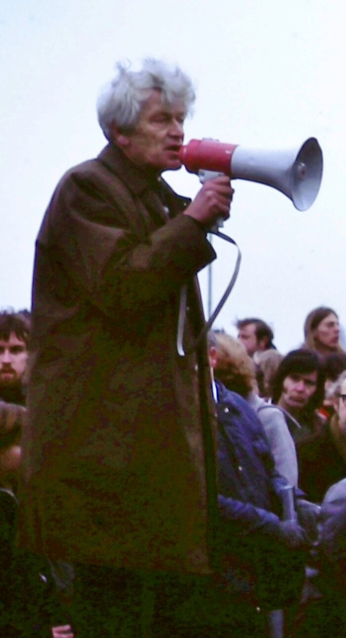 A white-haired man in a long coat speaking into a red megaphone in the open air, a crowd of listeners below and behind him under a grey sky