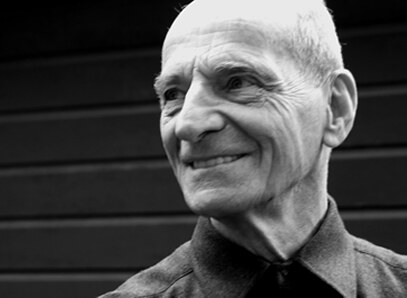 A black-and-white close portrait of an elderly man with a lined face, smiling slightly, looking off to one side, dark boards behind him