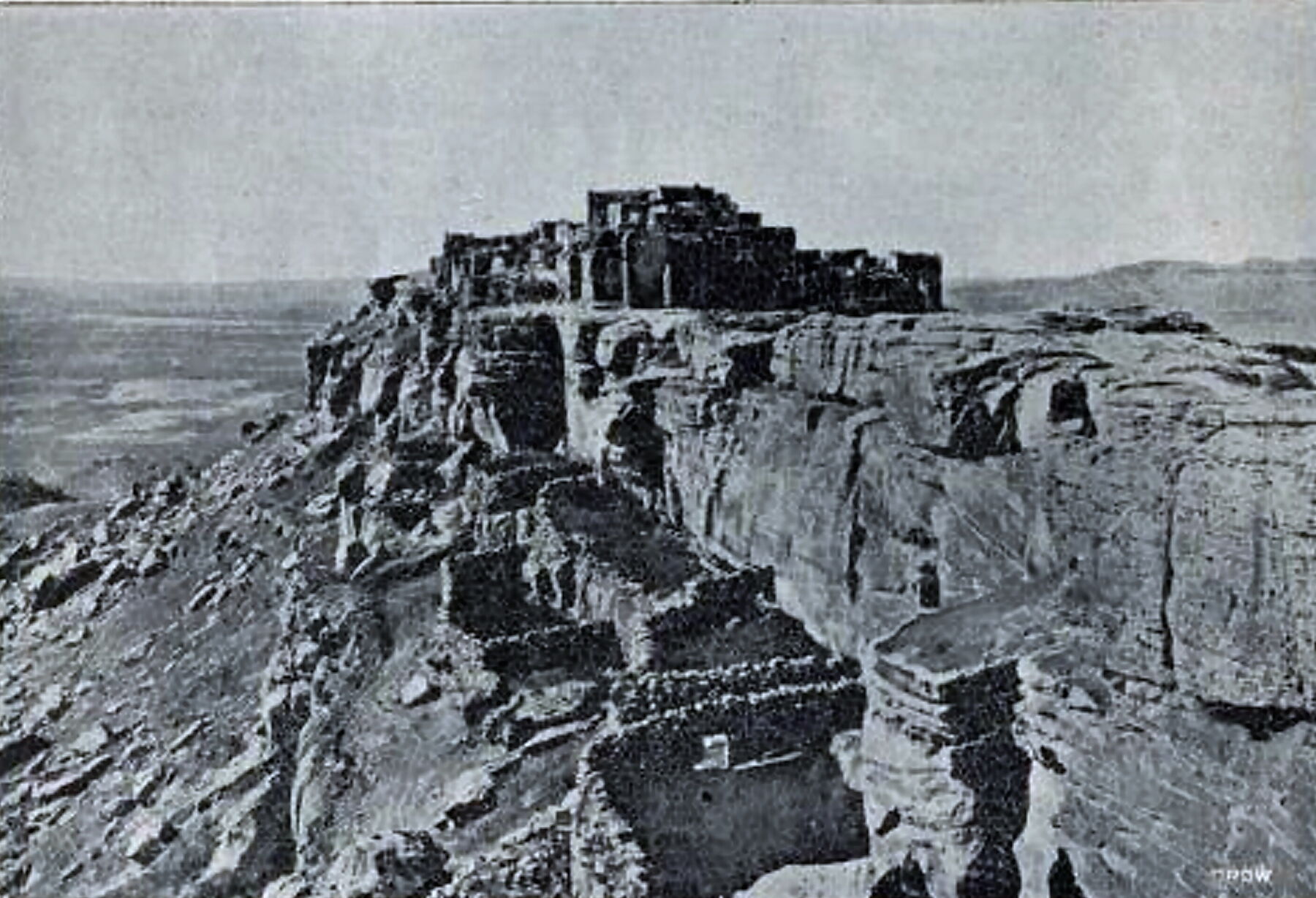 A black and white halftone of stone and adobe houses on the edge of a sheer-sided rock mesa, broken cliffs below and a wide plain beyond