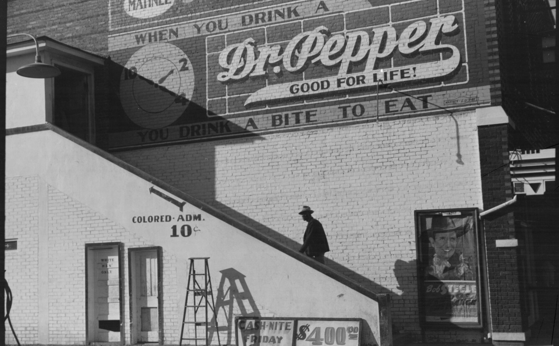 A black and white photograph of a brick movie theater: a man climbs an outside staircase along the side wall toward an upper door, beneath a sign reading Colored Adm. 10 cents, with a large Dr Pepper advertisement painted above