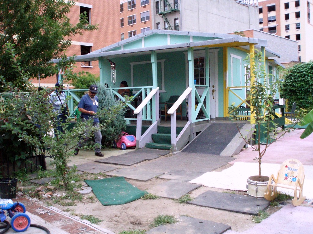 A small turquoise wooden house with a yellow-trimmed porch, railings and a ramp, in a garden with a young tree and paving stones, with brick apartment buildings rising behind it