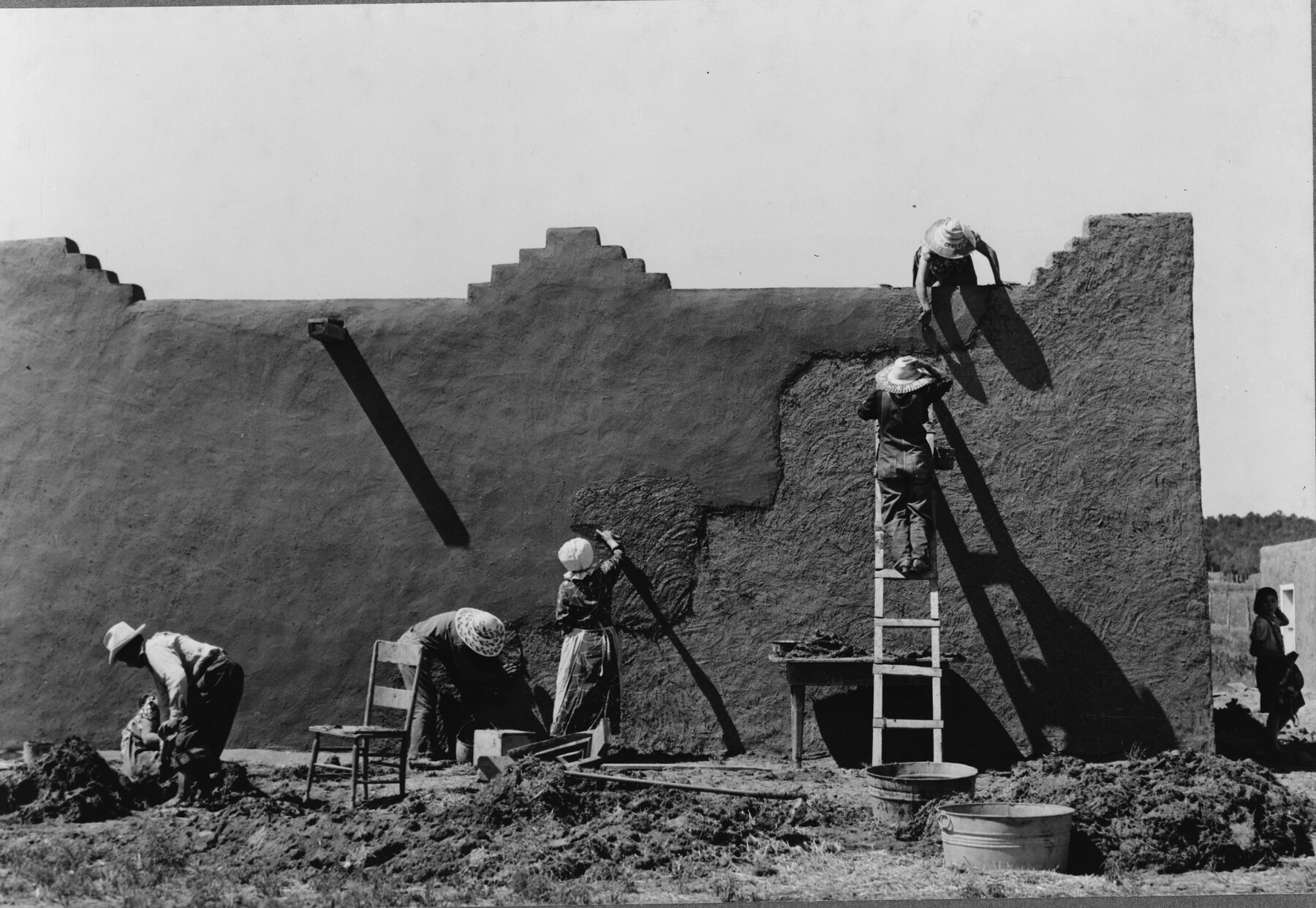 Several women in hats and long skirts replastering the mud wall of a flat-roofed adobe house with a stepped parapet; one stands on a ladder against the wall, another kneels at the top, and piles of mud, a tub and a chair sit in the foreground