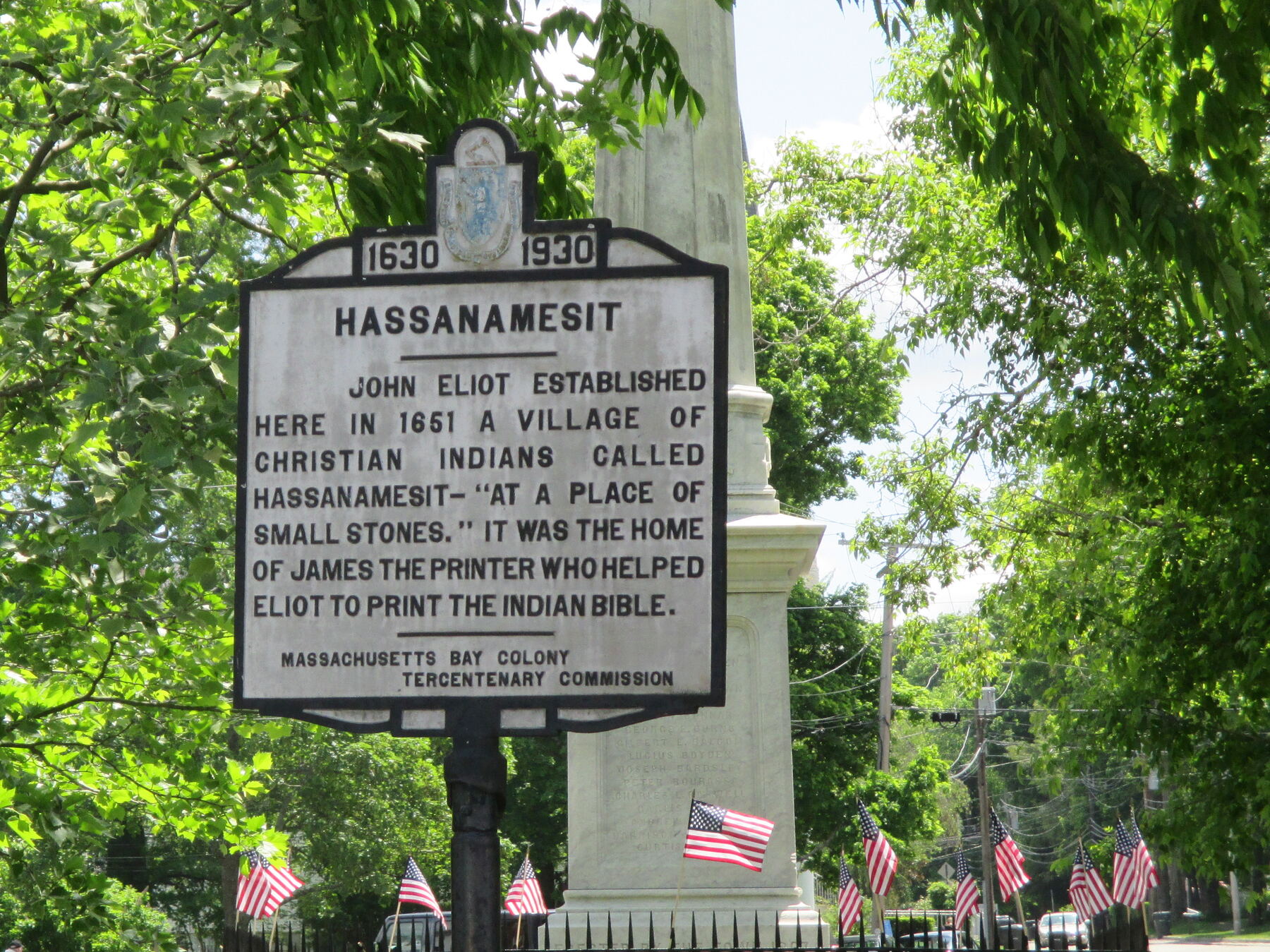 A white cast-metal roadside marker headed 1630–1930 under the Massachusetts seal, titled Hassanamesit, on a black post in front of a stone obelisk, with small American flags along a fence and trees behind