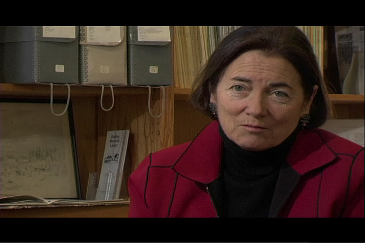A video still of a woman with short dark hair in a red jacket over a black turtleneck, speaking in front of shelves of archive boxes and papers