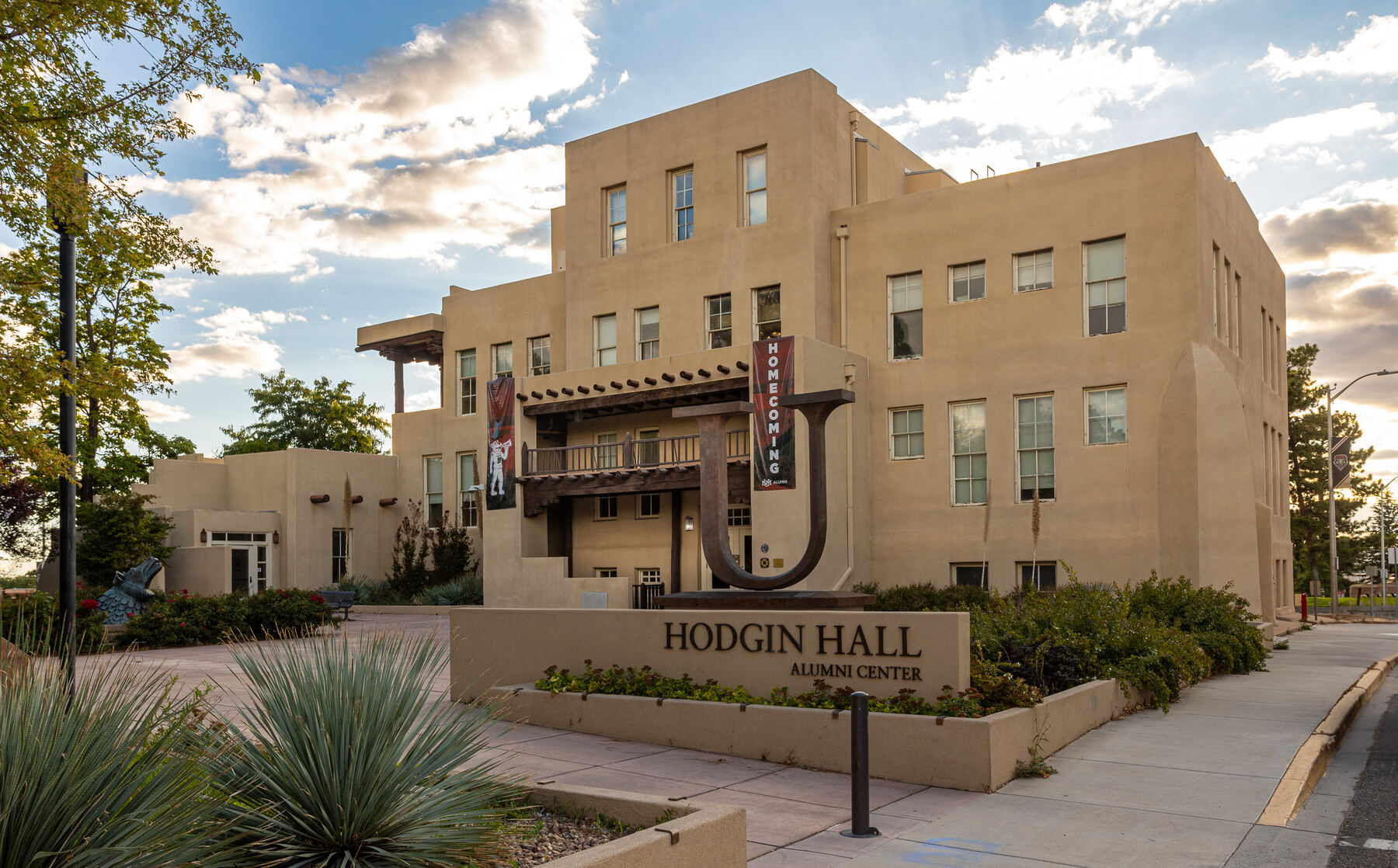 A flat-roofed, tan stucco building with stepped massing, projecting wooden beams and a wooden balcony, behind desert plants and a sign reading Hodgin Hall