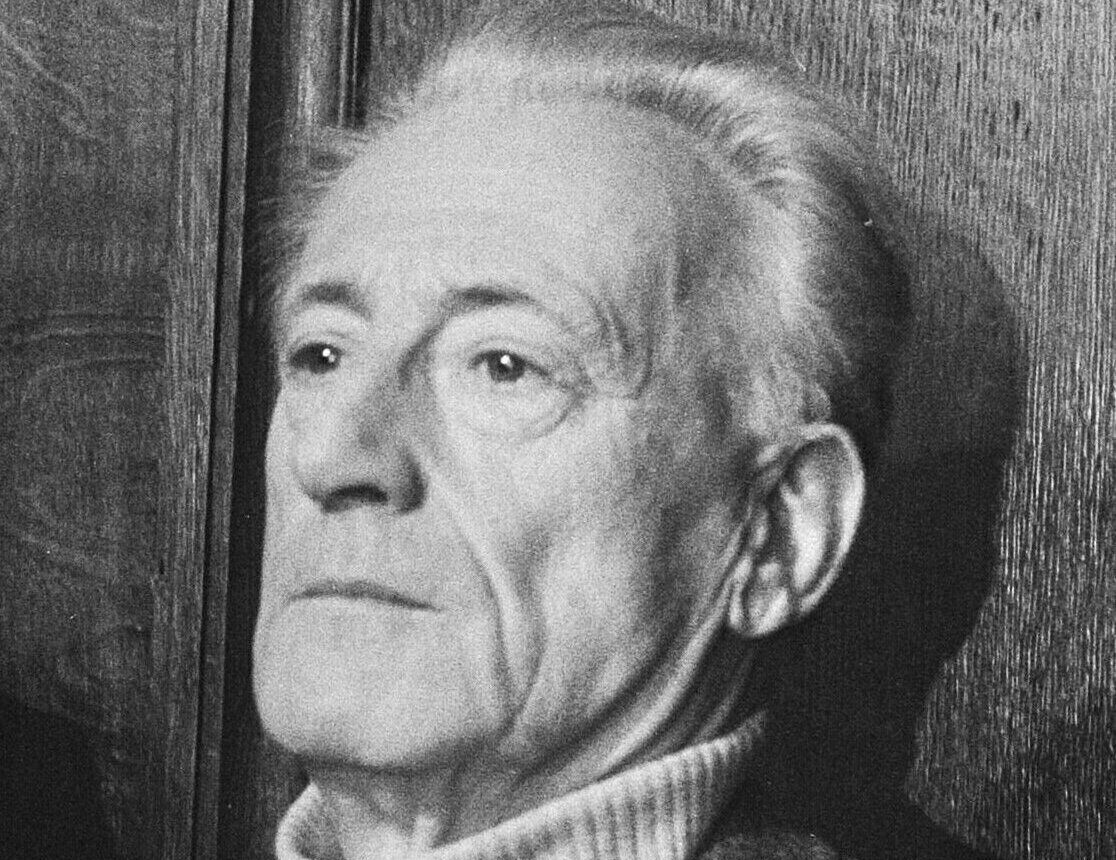 A black and white photograph of an older man with swept-back gray hair, in a jacket over a white turtleneck, looking to the side