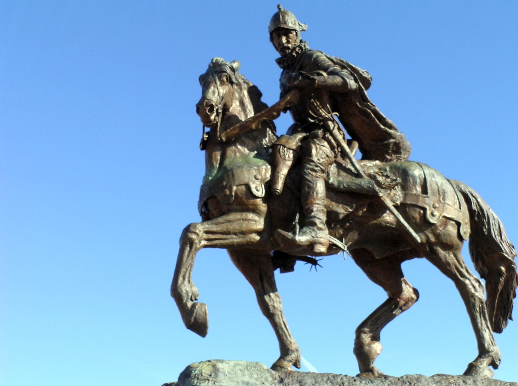 A bronze equestrian statue of a conquistador in a helmet and armor, holding a lance across his horse, which rears one foreleg, against a clear blue sky