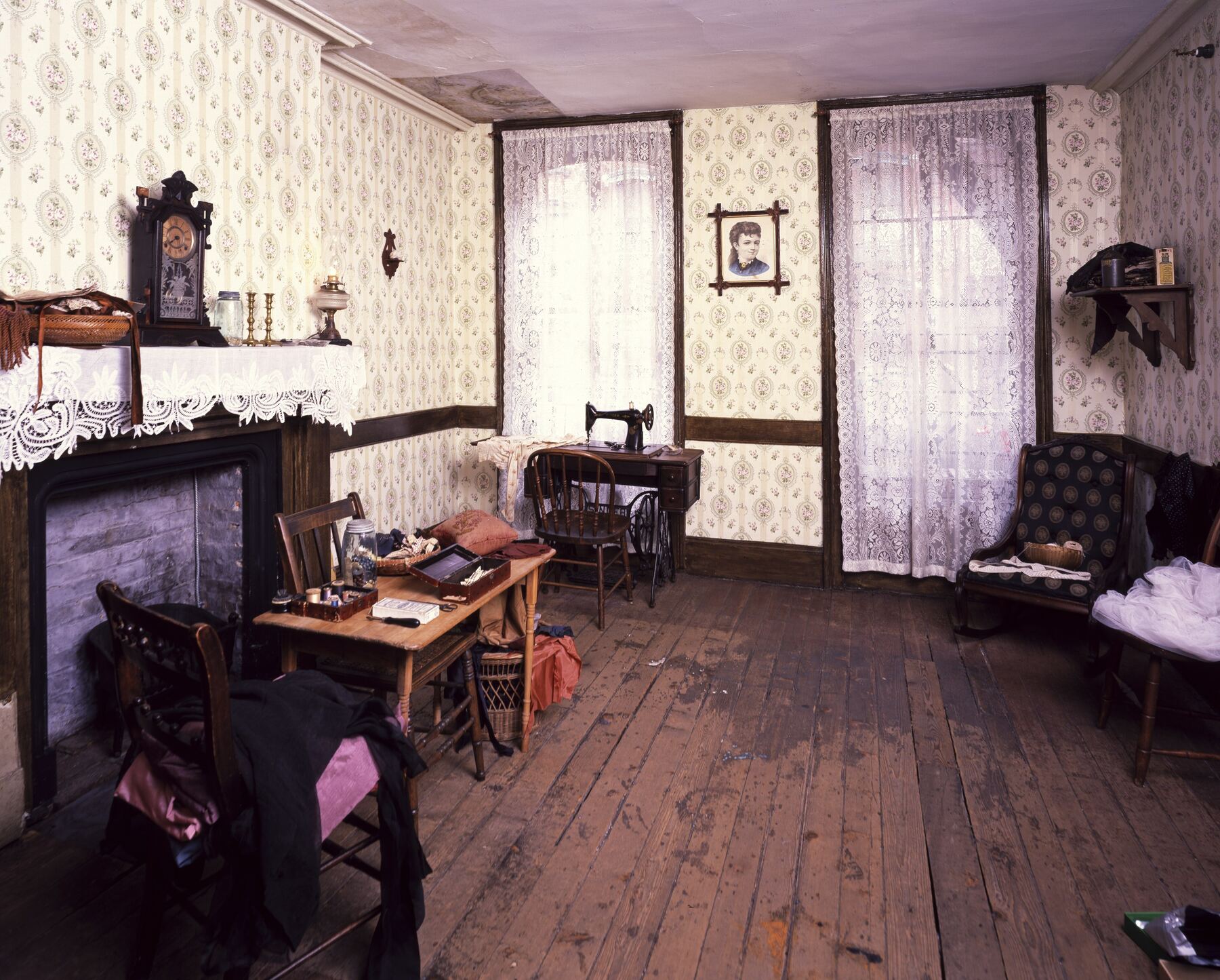 A restored nineteenth-century tenement parlor with patterned wallpaper, lace curtains, a treadle sewing machine by the window, a mantel clock, a small table and worn floorboards