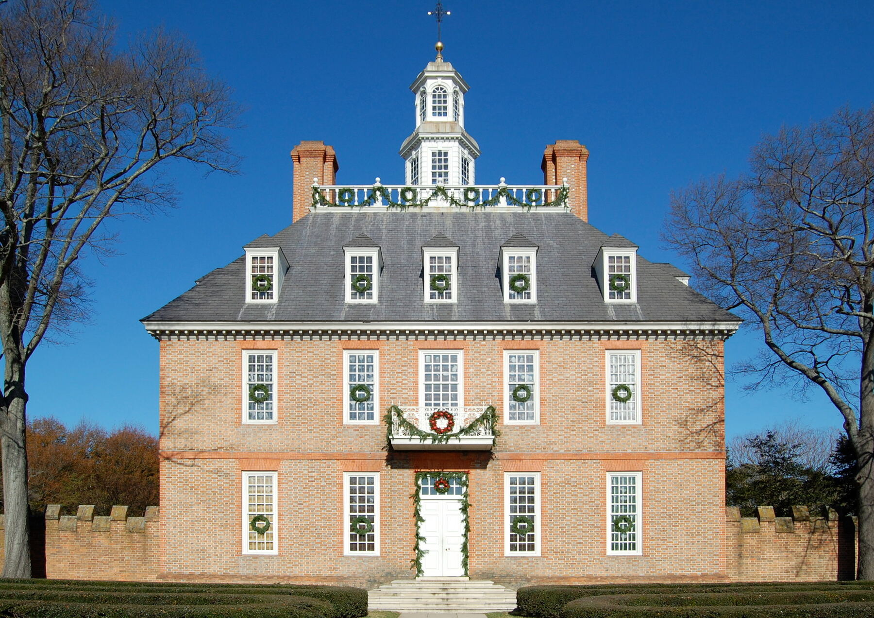 A symmetrical red-brick Georgian mansion with a steep slate roof, tall chimneys and a white cupola, behind a brick wall and wrought-iron gate under a blue sky