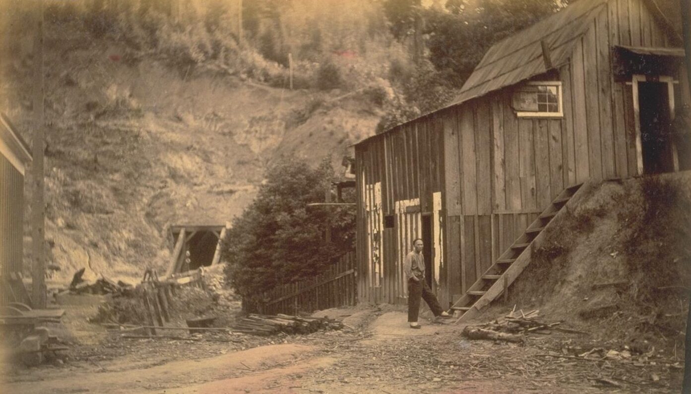 A sepia photograph of a board-and-batten building with an outside stair on a hillside, a Chinese man in a work shirt standing at the foot of the stair, and across a dirt yard a small timbered opening in a brushy slope, with lumber piled in front