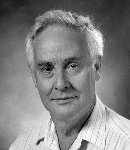 A black and white studio portrait of a man in late middle age with thick white hair, in a striped open-collared shirt, smiling slightly