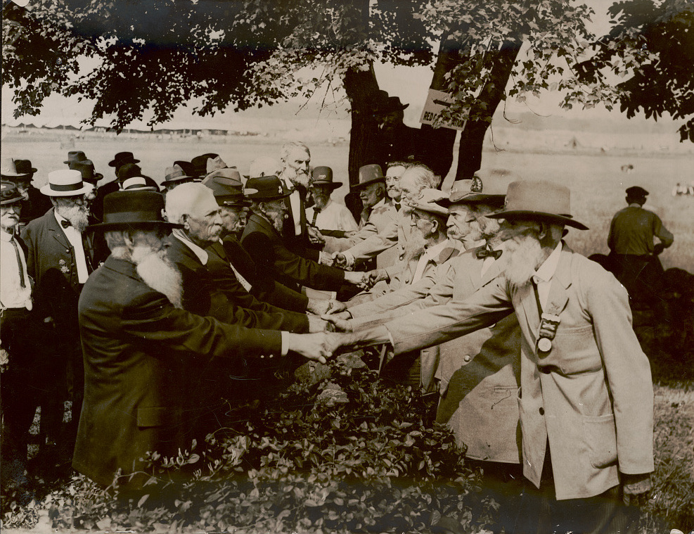 A sepia photograph: two lines of elderly bearded men in hats and suits reach across a low stone wall under trees to shake hands, with tents on the field behind them