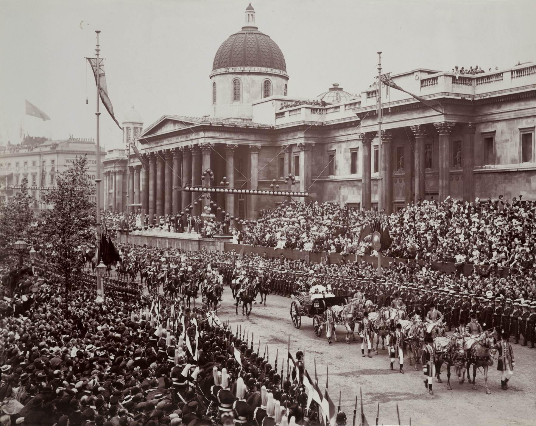 A sepia photograph: an open royal carriage drawn by plumed horses and flanked by footmen passes the domed National Gallery, between lines of soldiers and cavalry, with packed stands of spectators behind