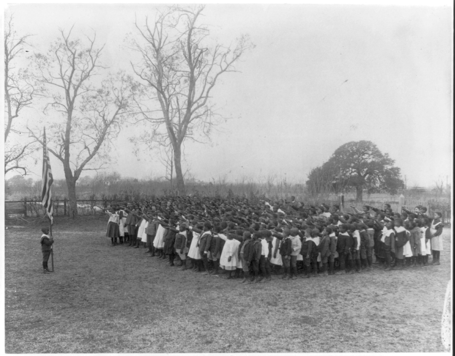 A black and white photograph: rows of Black schoolchildren on a bare field, right arms stretched out toward an American flag held by a small boy at the left, with leafless trees behind