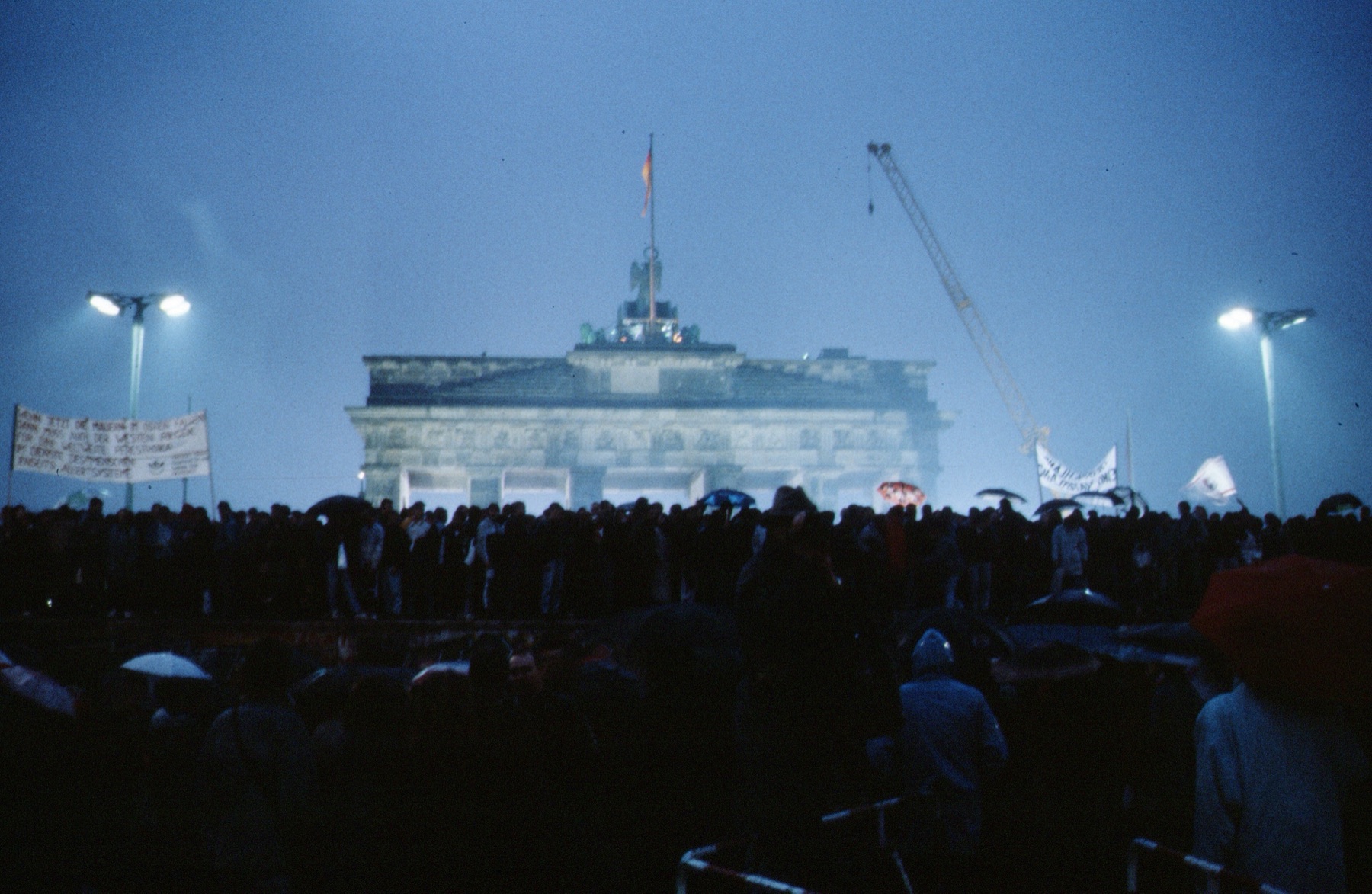 A color photograph at dusk: a dense crowd with umbrellas and banners stands before the Berlin Wall, with the floodlit Brandenburg Gate and a construction crane behind it
