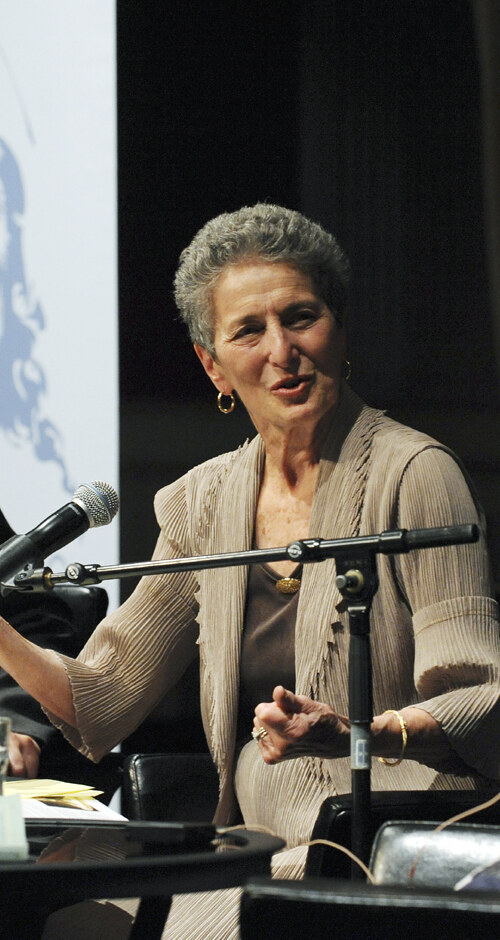 A color photograph of an older woman with short gray hair, in a pleated beige jacket, speaking and gesturing with both hands behind a microphone on a dark stage