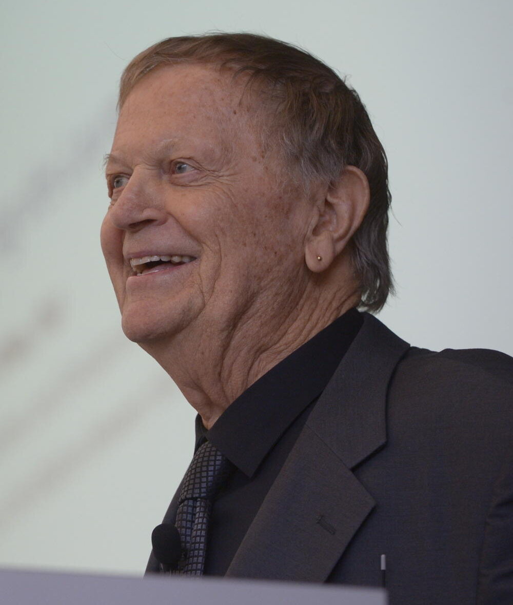 A color photograph of an elderly man in a dark suit and dark shirt, smiling broadly as he speaks at a lectern