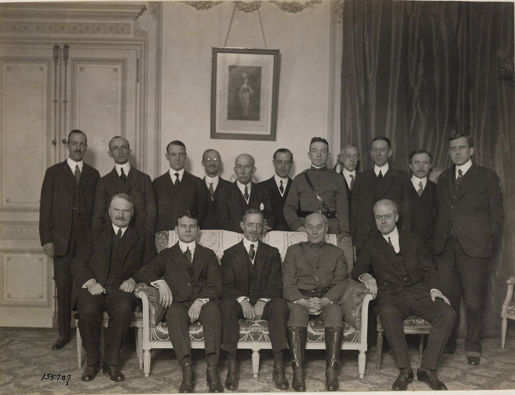 A sepia group photograph in a hotel salon: sixteen men in dark suits and two in army uniform, five seated on a sofa and chairs in front and eleven standing behind, under a framed portrait