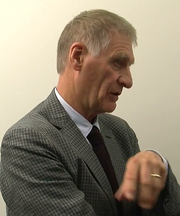 A video still of an older man with gray hair in a checked gray jacket and dark tie, looking to one side and gesturing as he talks