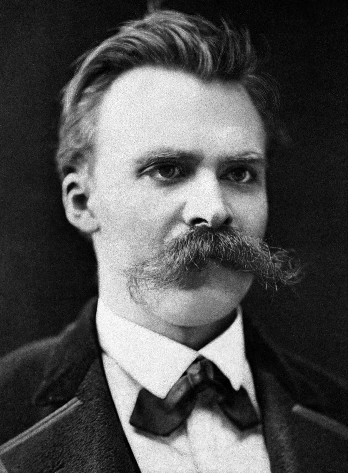 A black and white studio photograph of a young man with a heavy drooping mustache and swept-back hair, in a dark coat and a large bow tie