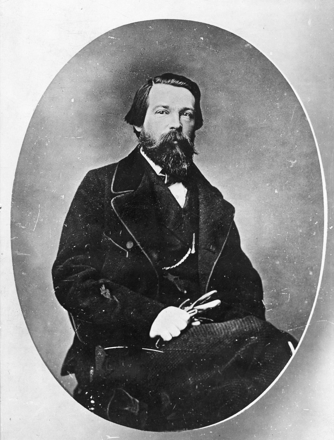 Oval-framed photograph of a seated man in his forties with dark hair, a pointed beard and moustache, in a dark coat and waistcoat, holding a pair of spectacles