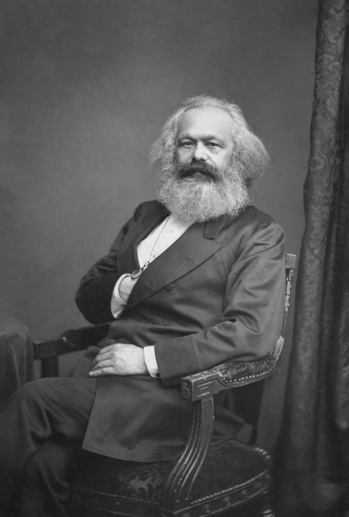 Studio photograph of a seated elderly man with a broad white beard and thick swept-back hair, arms folded across a dark double-breasted coat, a watch chain at his chest