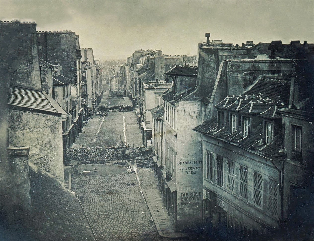An early photograph looking down a narrow Paris street of tall shuttered houses, empty of people, with two low barricades of piled paving stones blocking the roadway in the middle distance