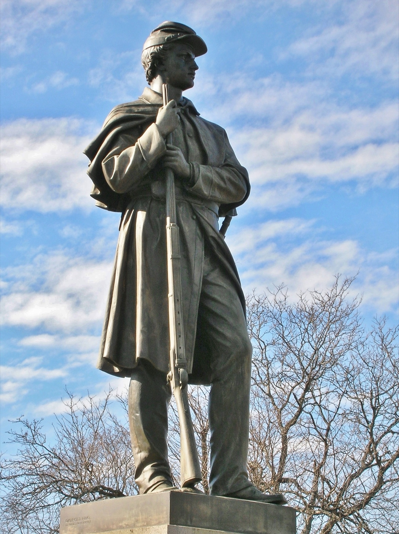 A bronze statue of a Civil War soldier in a kepi and long overcoat, both hands resting on the muzzle of a rifle, seen from below against a blue sky with thin cloud and bare branches