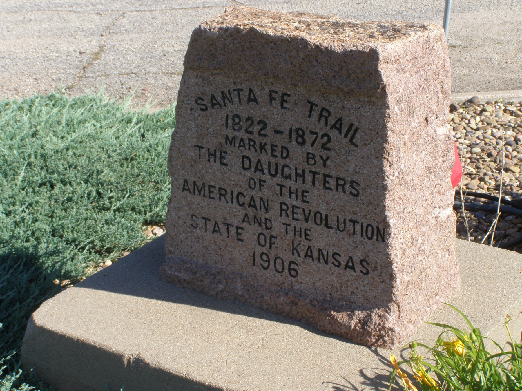 A pink granite marker on a concrete base, lettered in black: Santa Fe Trail, 1822–1872, marked by the Daughters of the American Revolution and the State of Kansas, 1906, with juniper and gravel beside it