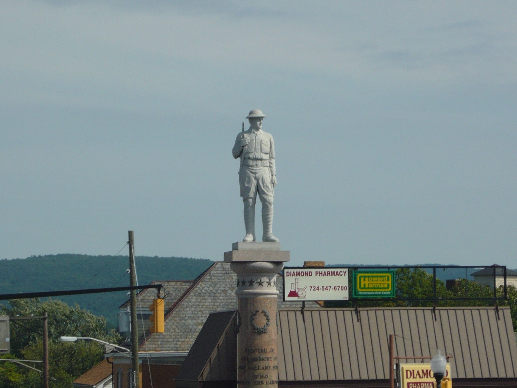 A pale stone statue of a World War I infantryman in a helmet, holding a rifle, on a tall column above shop roofs and a pharmacy sign, against a gray sky and wooded hills