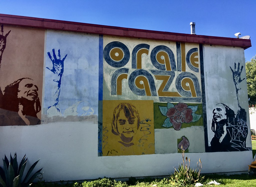 The end wall of a two-story stucco apartment building painted with a mural: the words Orale Raza in large outlined letters, a blue hand, painted faces, a rose, and a dark portrait of a man with long hair, under a blue sky