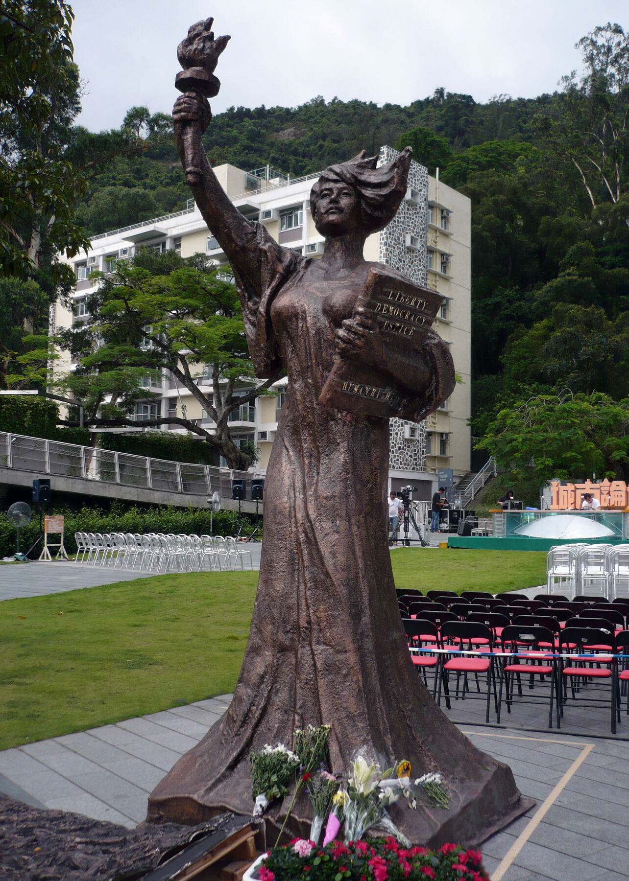 A bronze statue of a woman in a long robe holding a torch high in her right hand and a book in her left, on a lawn with rows of folding chairs set out before it and a university building and wooded hill behind