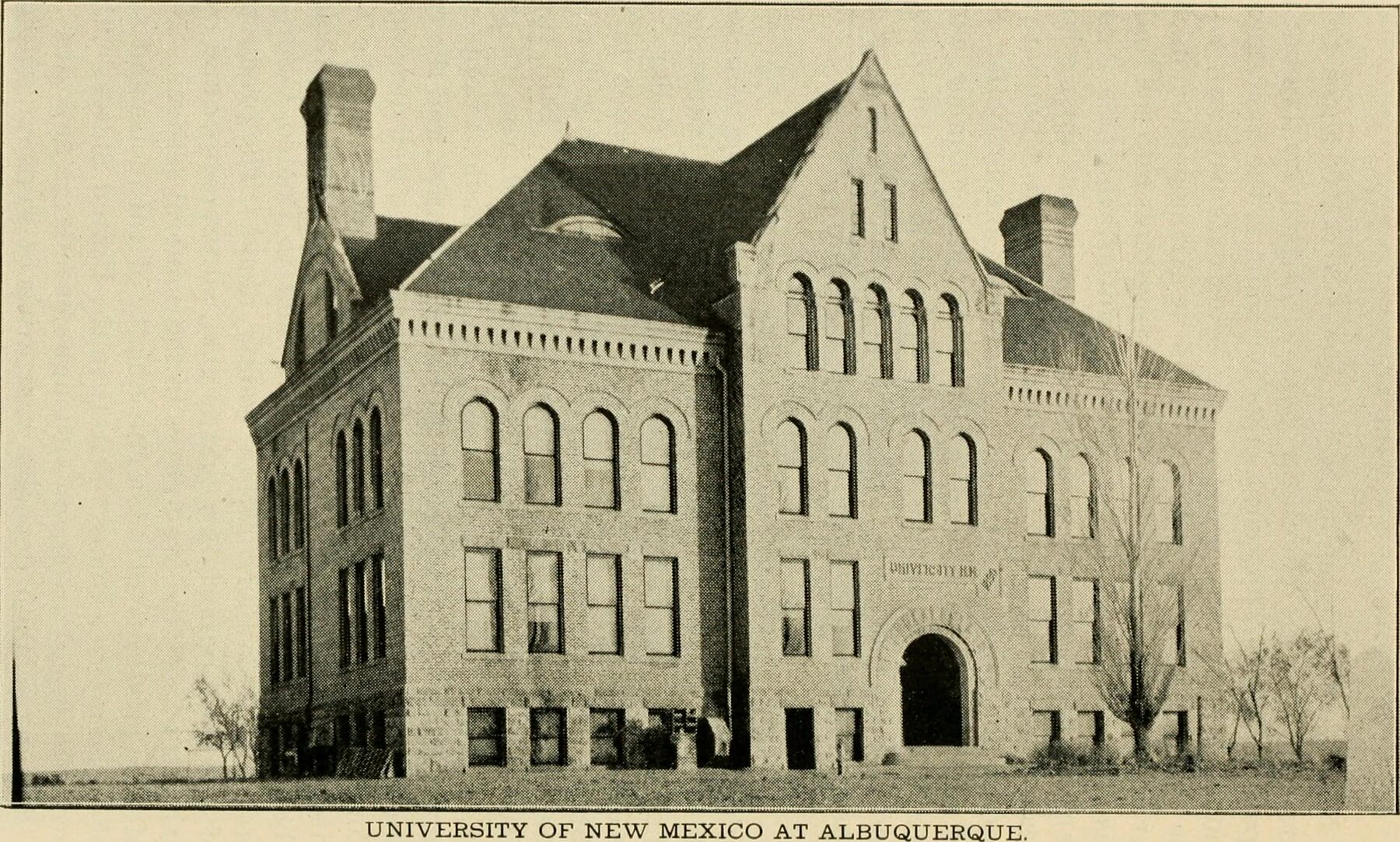 A printed photograph of a three-story brick building with steep gables, tall chimneys and arched windows, standing alone on bare ground
