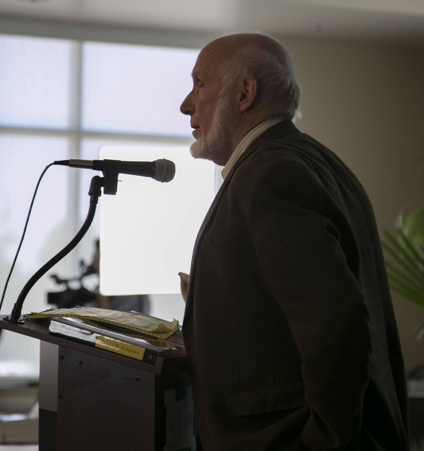 A color photograph of a bald, white-bearded man in a dark jacket, seen in profile, speaking into a microphone at a lectern