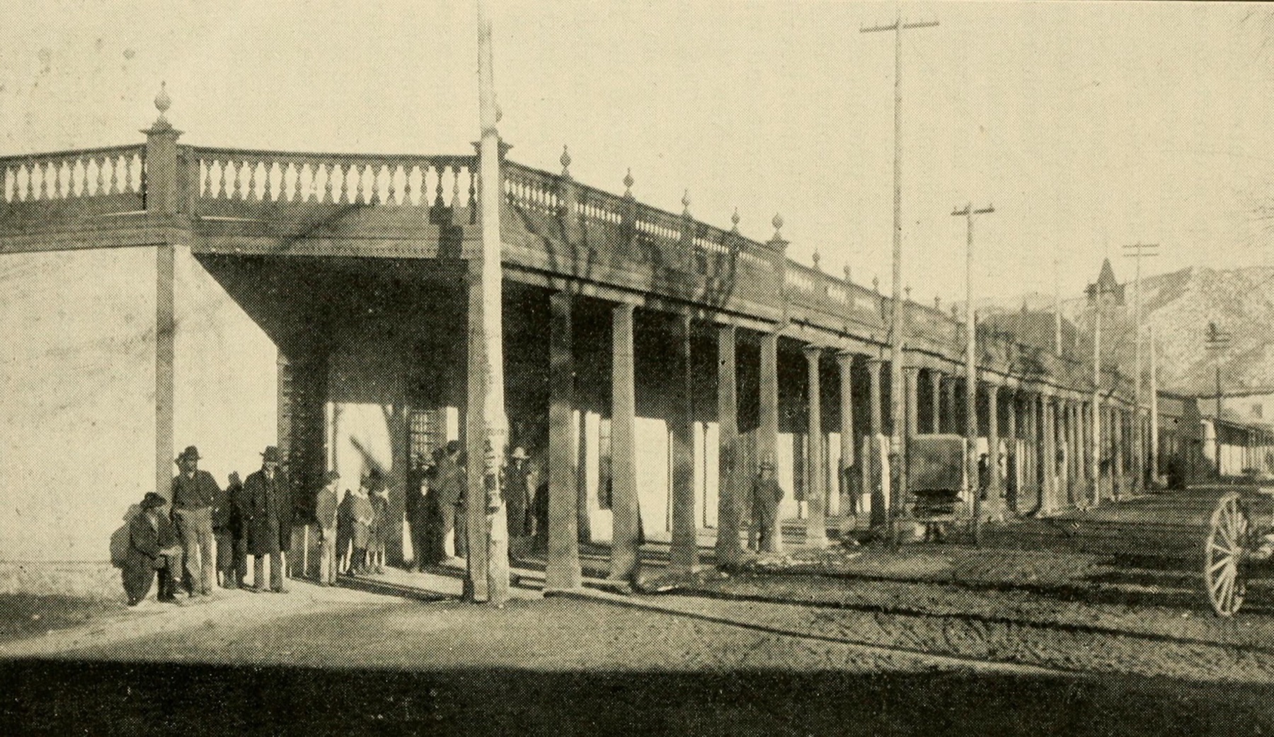 A black and white halftone of a long adobe building in Santa Fe with a deep porch of tall columns under a balustrade, men and boys standing in its shade, utility poles along the dirt street and a wagon wheel at the edge of the frame
