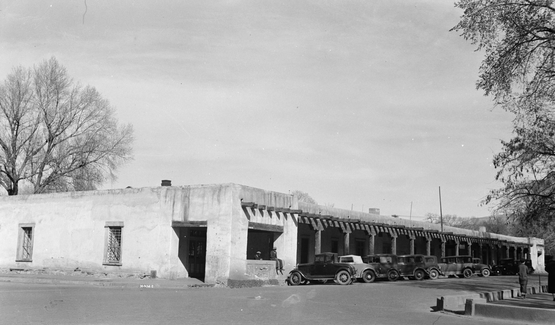 A black and white photograph of a long, flat-roofed adobe building with plain plastered walls and a porch of rough posts under projecting beams, with several 1930s cars parked along the front and bare trees at either side