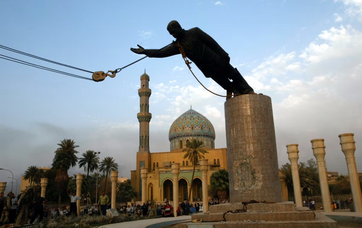 A bronze statue of a standing man tilts from its plinth, hauled by cables, with a domed building and palms behind and a thinly populated plaza below